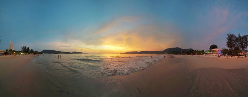 Wide beach panorama at sunset in Phuket, Thailand showing ocean waves, sandy shore, scattered tourists, distant mountains, and golden sky with clouds