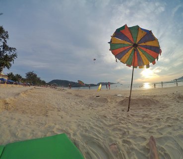 Colorful striped beach umbrella on sandy beach at sunset with people walking along shoreline, ocean waves, distant hills, and parasailers in the sky over Phuket, Thailand