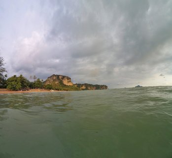Coastal seascape showing limestone cliffs and karsts along a tropical beach in Ao Nang, Thailand, with overcast cloudy sky and green-tinted ocean water in the foreground