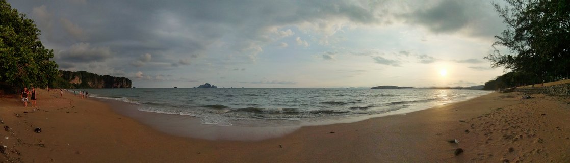 Panoramic beach scene at sunset showing sandy shore, ocean waves, limestone cliffs on the left, distant islands, and tropical trees framing both sides with sun setting on the right