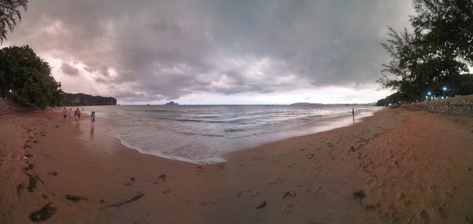 Wide beach scene at Ao Nang showing sand, ocean waves, cloudy sky, scattered people walking, trees on both sides, and distant islands on the horizon during evening twilight