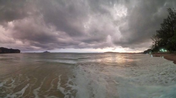 Wide beach scene showing ocean waves washing onto sand under heavy gray storm clouds with sunset light breaking through on the horizon, trees visible on the right side and distant islands on the left