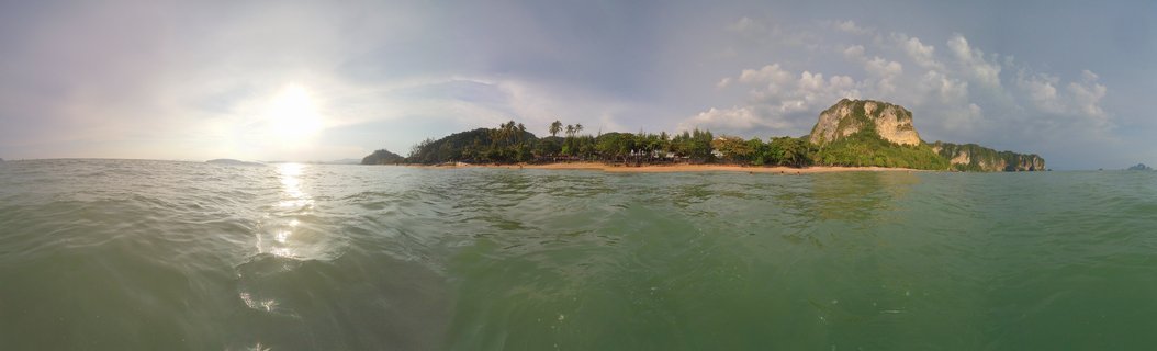 Panoramic seascape showing tropical island with limestone cliffs, palm trees, and sandy beach surrounded by turquoise water under partly cloudy sky in Ao Nang, Thailand