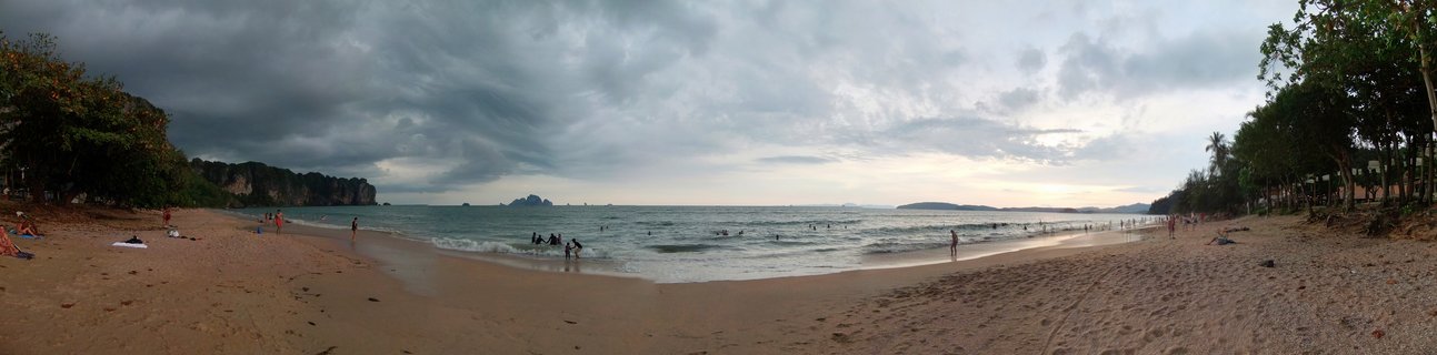 Panoramic beach scene showing Ao Nang in Thailand with people on sand and in water, tropical trees on both sides, limestone cliffs in distance, dramatic cloudy gray sky overhead