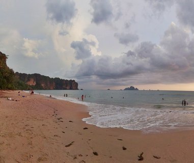 Wide beach scene showing golden sand with footprints, gentle waves washing ashore, people swimming in turquoise water, and dramatic limestone cliffs covered in vegetation on the left under a cloudy sky at sunset