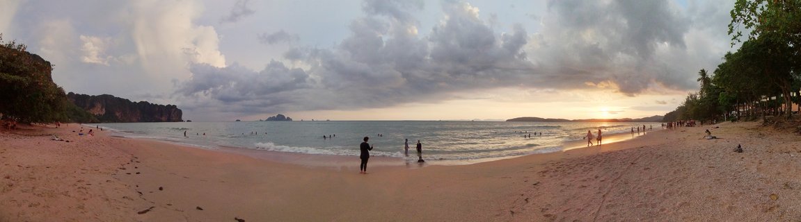 Panoramic beach sunset scene showing silhouetted people on sand and in ocean water, with limestone cliff formations on the left and tropical trees framing both sides under cloudy sky with setting sun