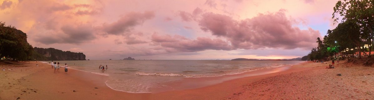 Wide panoramic beach scene at sunset showing golden sand, ocean waves, people walking on the shore, limestone cliffs on the left, tropical trees, pink and purple clouds filling the sky, and distant islands on the horizon