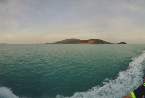 View from a moving boat showing turquoise ocean water with white wake, tropical forested islands in the distance under a pale sky with visible moon