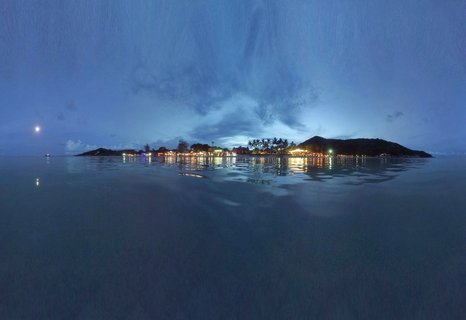 Panoramic coastal view of Koh Phangan island at twilight with illuminated beachfront resorts and restaurants reflecting on calm gulf waters under a blue evening sky with visible moon