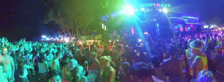 Large crowd of people at a beach party at night with neon green and blue lighting, palm trees, and a bar building with illuminated signage in the background
