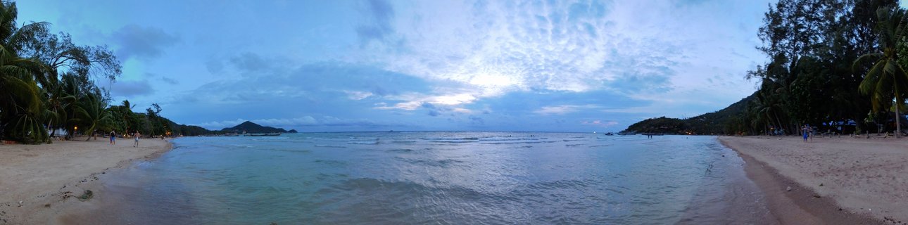 Wide panoramic photograph of a tropical beach bay at dusk showing sandy shores on both sides with palm trees, calm ocean water in the center, and forested hills in the distance under a cloudy blue sky
