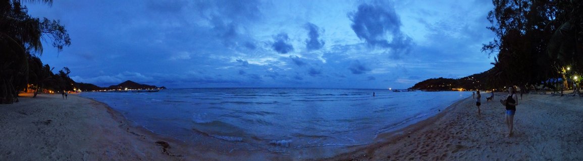 Panoramic beach scene at dusk showing curved bay with sand beach on both sides, calm ocean water in center, palm trees, distant hills with lights, people walking on beach, blue cloudy sky
