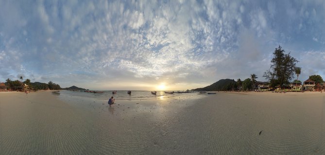 Panoramic beach scene at sunset in Koh Tao, Thailand, showing sandy shore, multiple longtail boats in shallow water, person on beach, tropical trees, hills, and cloudy sky with golden sunlight