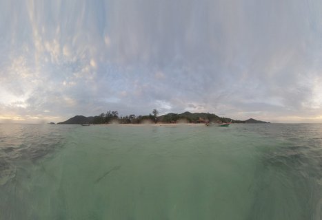 Panoramic tropical seascape showing turquoise ocean water in foreground with a Thai island featuring beach resorts, palm trees, hills, and traditional long-tail boats under cloudy sky