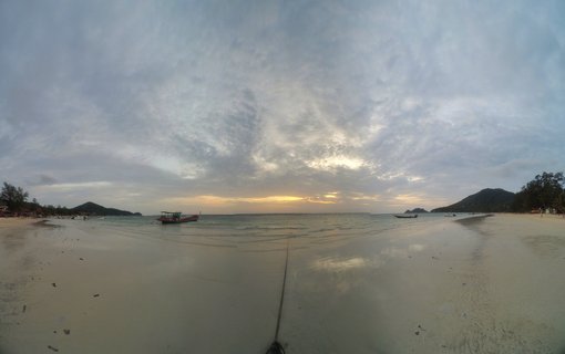 Panoramic view of a tropical beach in Koh Tao, Thailand with traditional long-tail boats in calm water during sunset, sandy shore in foreground with hills and cloudy sky