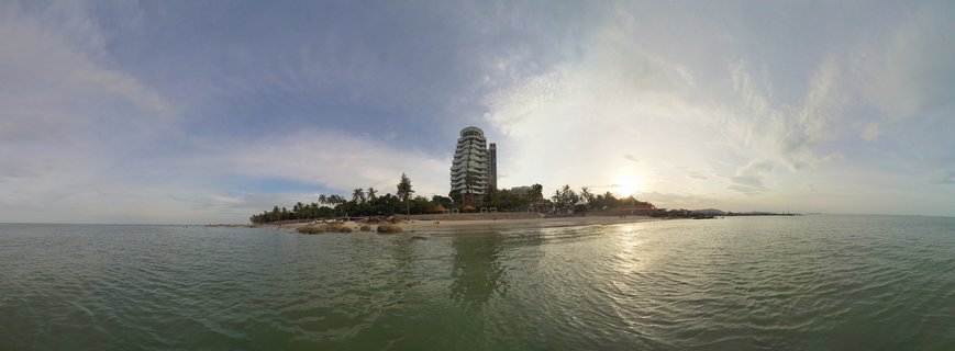 Panoramic view of a tall white resort hotel on a tropical beach in Hua Hin, Thailand, with palm trees, calm ocean water, and sunset sky with clouds