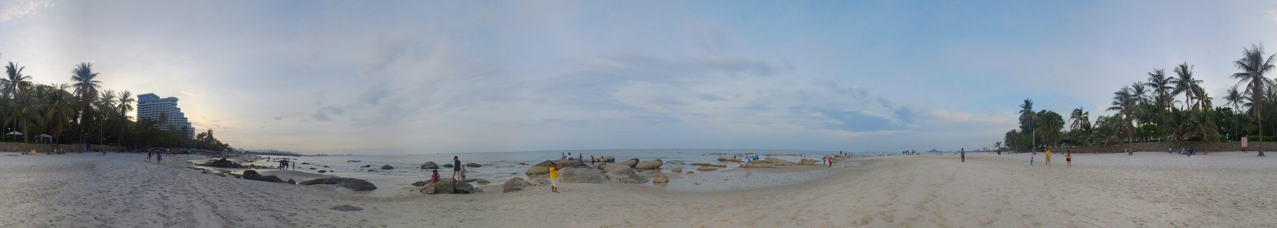 Wide panoramic beach scene showing sandy shore with scattered people, large rocks in shallow water, palm trees on both sides, ocean horizon, and a hotel building in the distance under a partly cloudy sky
