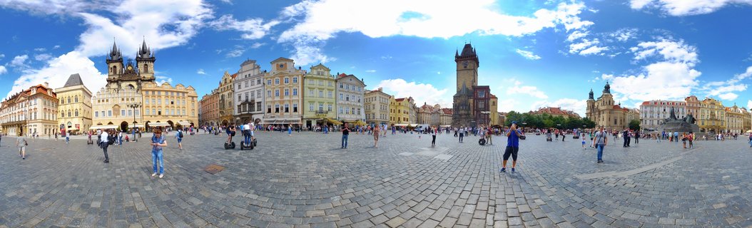 Wide panoramic view of Prague's Old Town Square showing cobblestone pavement, tourists walking, colorful historic buildings, the twin-spired Týn Church, and the Old Town Hall Tower under a blue sky with scattered clouds