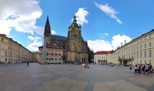St. Vitus Cathedral with Gothic spire and green copper roof at Prague Castle courtyard with tourists on paved plaza under blue sky with clouds