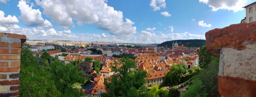 Panoramic cityscape of Prague showing dense clusters of buildings with orange-red tiled roofs, green trees, church spires, and a blue sky with white clouds, viewed from an elevated castle position