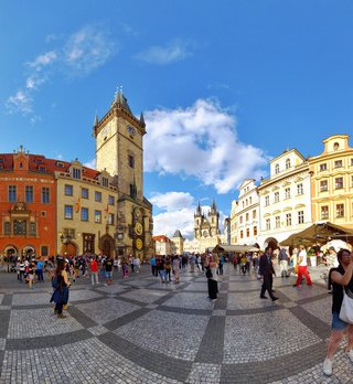 Wide view of Prague's Old Town Square showing the Astronomical Clock tower, colorful historic buildings, cobblestone pavement, and tourists walking under a blue sky with white clouds