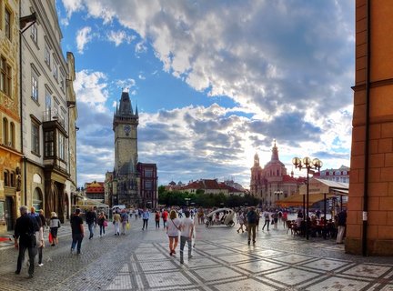 Wide view of Prague Old Town Square showing the Astronomical Clock tower, Church of Saint Nicholas, historic buildings, and pedestrians on cobblestone pavement under cloudy sky at sunset
