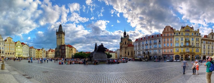 Wide panoramic view of Prague's Old Town Square showing the Astronomical Clock tower, Jan Hus Memorial statue, colorful historic buildings, and tourists on cobblestone pavement under cloudy blue sky