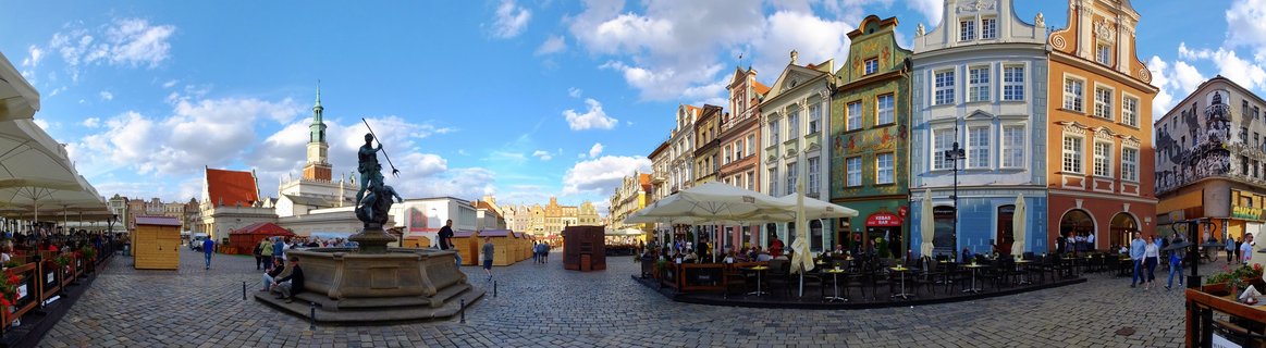 Wide view of Poznan's Old Market Square showing colorful Renaissance buildings with ornate gables, a bronze fountain statue in the foreground, cobblestone pavement, outdoor restaurant seating with white umbrellas, and blue sky with scattered clouds