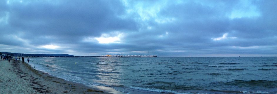 Panoramic view of illuminated Sopot Pier extending into Baltic Sea at dusk with people walking on sandy beach under cloudy sky