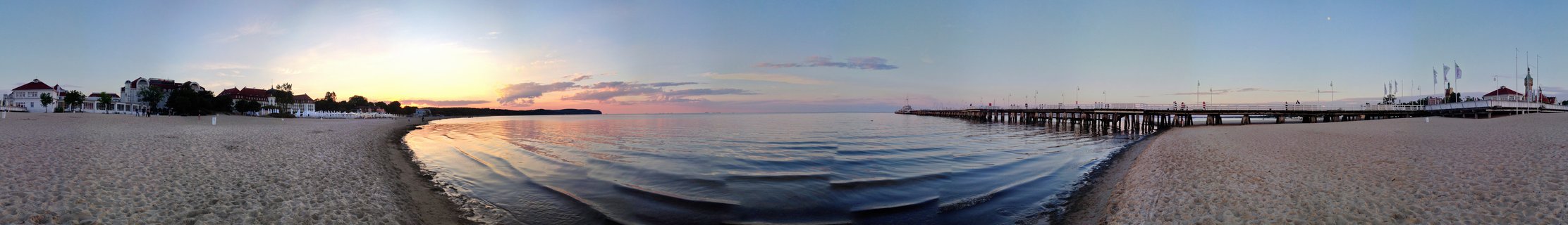 Panoramic view of Sopot Pier extending into the Baltic Sea at sunset with sandy beach, calm waters, and coastal buildings under a colorful sky