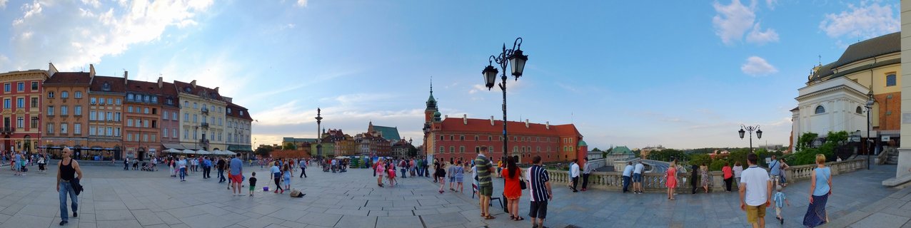 Wide panoramic view of Warsaw Old Town Market Square showing colorful historic buildings, tourists walking on cobblestone plaza, ornate street lamps, and blue sky with scattered clouds