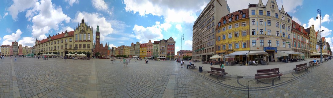 Wide panoramic view of Wroclaw Market Square showing historic buildings with colorful facades, Gothic Town Hall tower, cobblestone plaza with people walking, outdoor cafes, blue sky with white clouds
