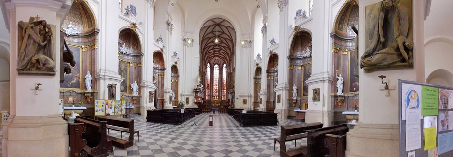 Wide interior view of a Baroque church showing a checkered black and white floor, rows of wooden pews, white walls with ornate side chapels containing religious artwork and statues, vaulted ceiling with wooden beams, and stained glass windows at the far end