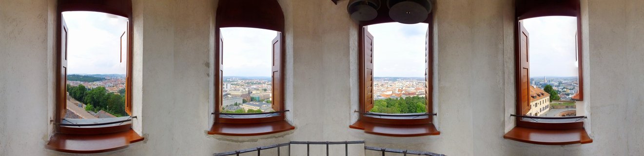 Interior view showing four arched windows with brown wooden frames and open shutters, each displaying a panoramic view of Brno city with red rooftops and green trees under cloudy sky