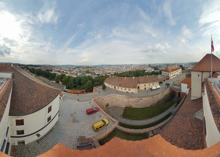 Aerial panoramic view of Špilberk Castle courtyard with historic buildings, red-tiled roofs, cobblestone paths, and Brno city visible in the background under blue sky with clouds.
