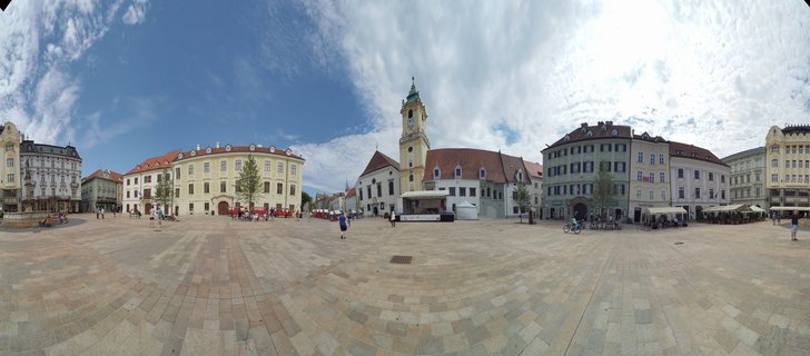 Wide panoramic photograph of a European town square showing historic buildings with a prominent church tower, paved plaza with pedestrians, outdoor cafes, and blue sky with white clouds
