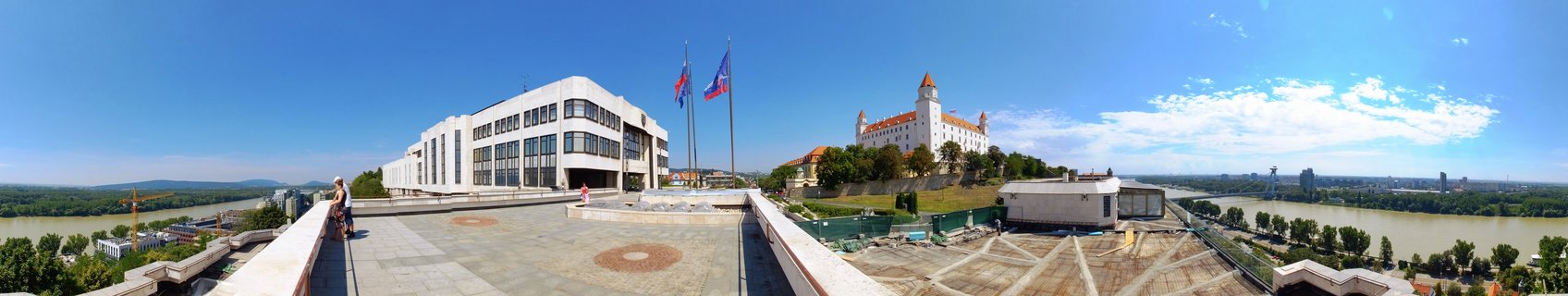 Panoramic view of Bratislava Castle, a white medieval fortress with four red-tiled corner towers on a hilltop, overlooking the Danube River. The modern white National Council building stands nearby with Slovak and European Union flags. Clear blue sky, summer day, viewing terrace with visitors, urban landscape and green trees.