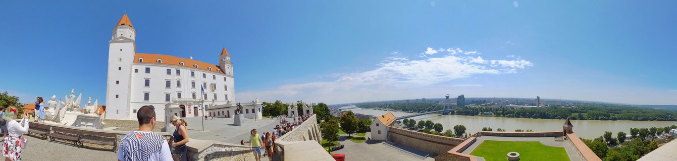 White Bratislava Castle with orange-tiled towers on a hill overlooking the Danube River, with tourists walking on the castle grounds under a blue sky