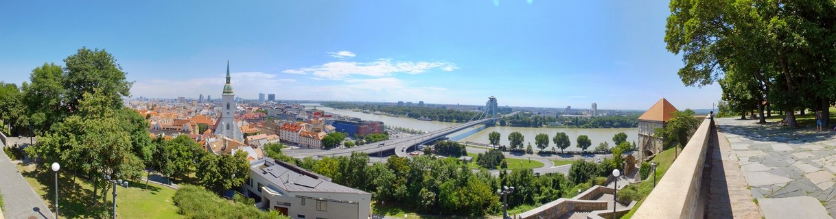Panoramic cityscape of Bratislava showing the Danube River, UFO Bridge with its distinctive tower, St. Martin's Cathedral with green spire, red-roofed Old Town buildings, and lush green trees under a clear blue sky