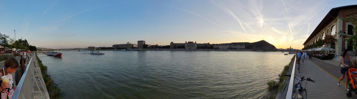 Wide panoramic photograph of the Danube River in Budapest at sunset showing Buda Castle buildings and Gellért Hill across the water, with boats on the river and people walking along the riverside promenade