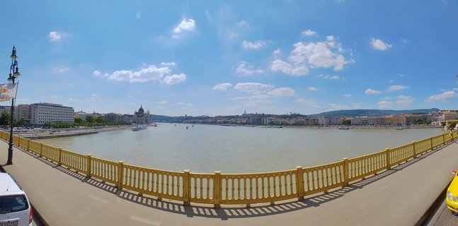 Wide panoramic view of Budapest's Danube River with yellow bridge railing in foreground, Hungarian Parliament Building visible on the left bank, and Buda Castle hills in the distance under blue sky with scattered clouds