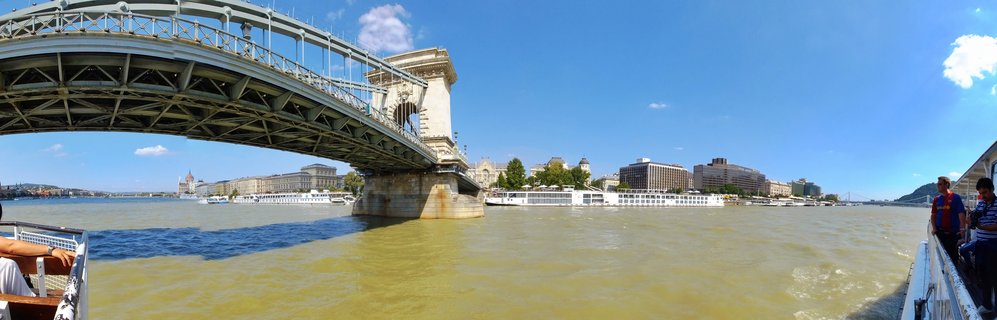 View of Budapest's Chain Bridge over the Danube River from a boat, with the Parliament Building visible in the distance and river cruise ships along the waterfront under a blue sky