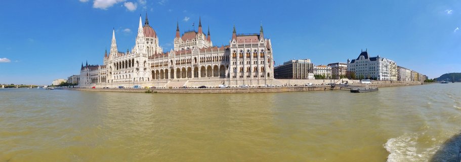 Wide panoramic view of the Hungarian Parliament Building along the Danube River under a clear blue sky with scattered white clouds