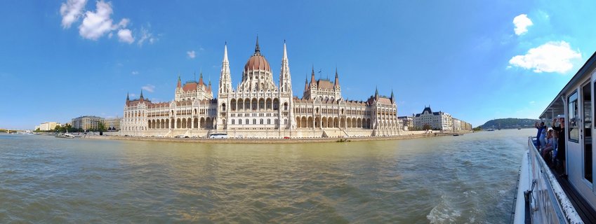 Wide view of the Hungarian Parliament Building from the Danube River showing the Gothic Revival architecture with central dome and spires under blue sky with tourists visible on a cruise boat