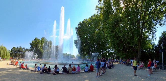 Large decorative fountain with multiple water jets in a public park, surrounded by people sitting on the fountain's edge under mature trees on a sunny day