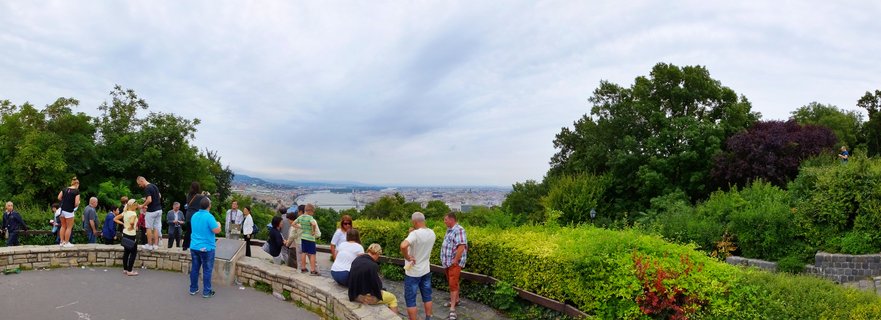 Wide panoramic view showing tourists at a hilltop viewpoint overlooking Budapest city and the Danube River, with green trees framing the scene on both sides under cloudy sky