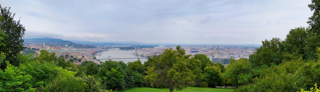 Wide panoramic photograph of Budapest showing the Danube River with bridges, Parliament Building, city buildings, and green trees in the foreground under cloudy sky