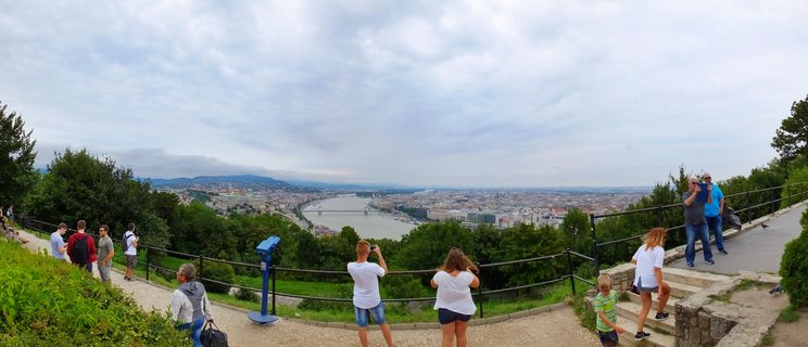 Panoramic view of Budapest city and Danube River from elevated viewpoint with tourists taking photographs along a fenced overlook area with trees and greenery in the foreground