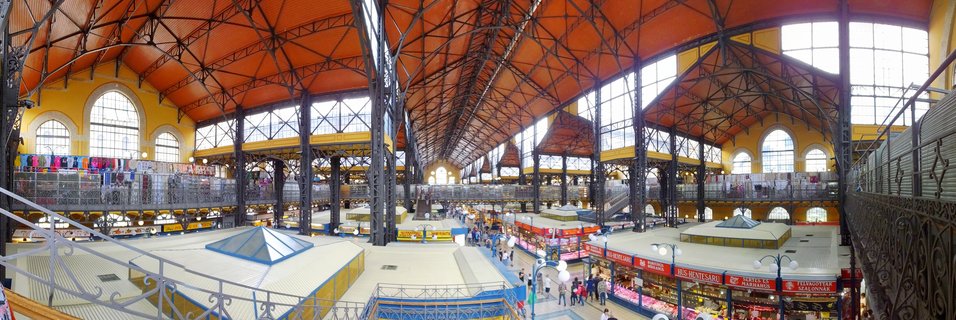 Interior panorama of Budapest's Great Market Hall showing ornate black iron columns, orange arched ceiling, yellow walls with arched windows, and multiple levels of market stalls with shoppers
