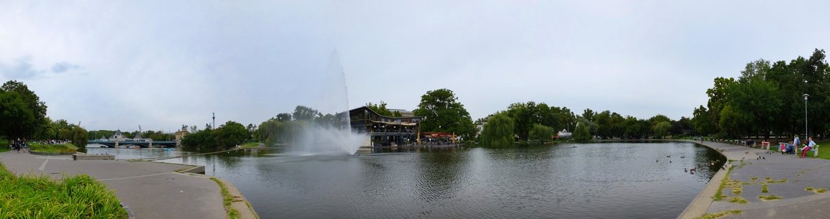 Wide panoramic view of a city park lake with a tall fountain spraying water, a wooden restaurant building on the shore, tree-lined walkways, and people strolling along the paths under an overcast sky.